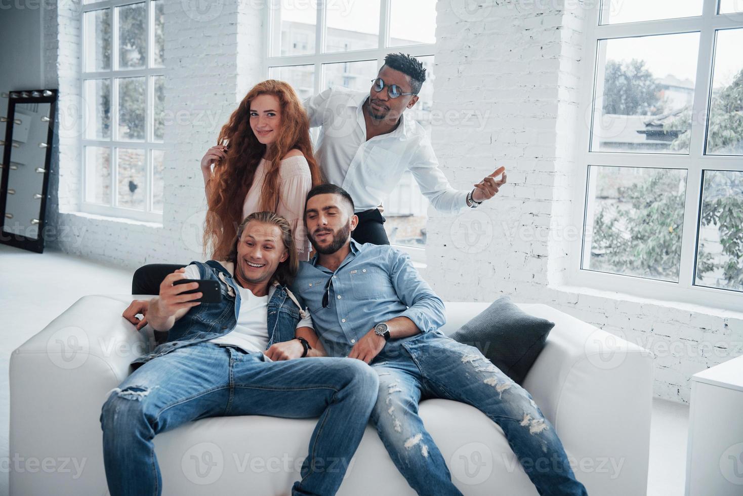 eine Vielzahl von Emotionen. Fröhliche junge Freunde, die Selfies auf Sofa und weißem Interieur machen foto