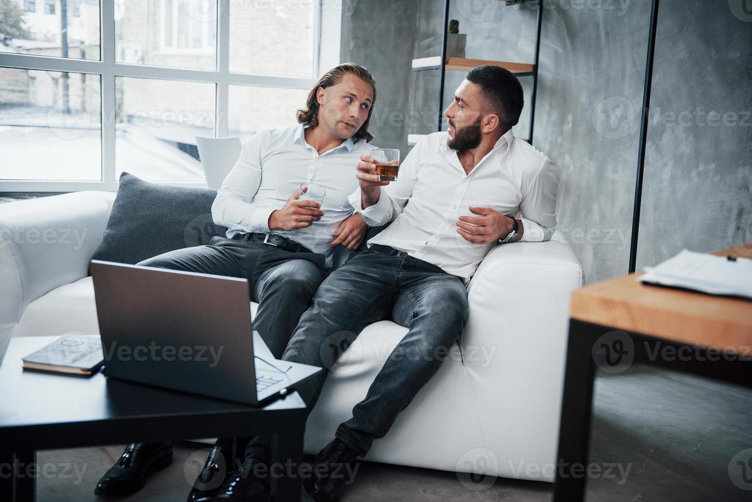 Zwei Geschäftsmann, die vor Laptop sitzen, über ihre Pläne sprechen und Whisky trinken foto