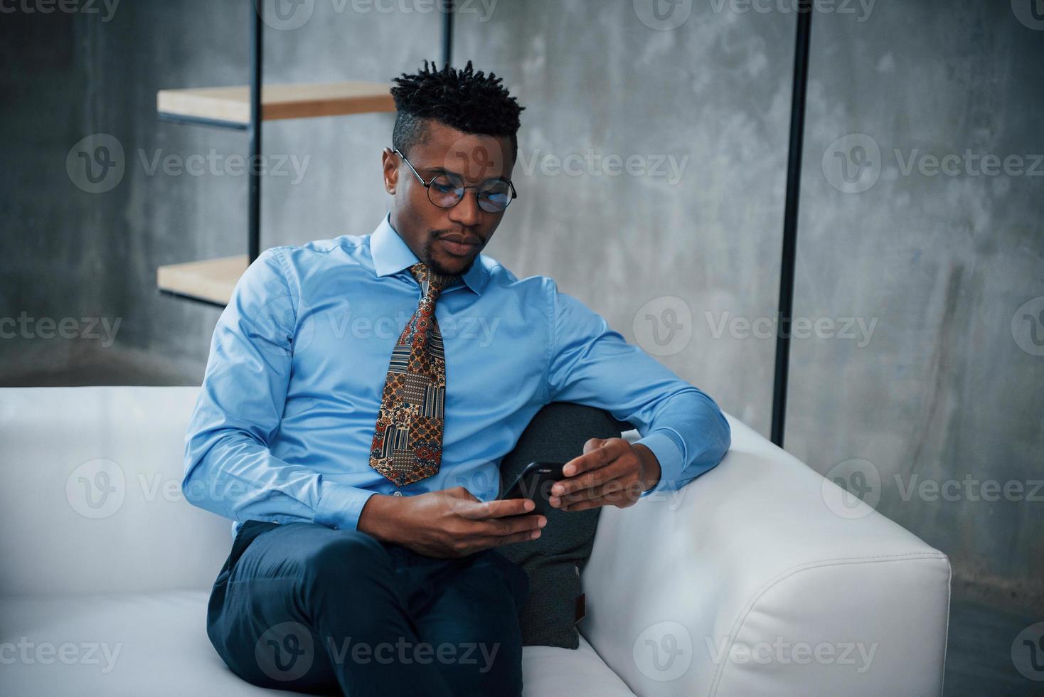 Nahaufnahme des Porträts eines jungen Afroamerikaners mit Brille und klassischer Kleidung, der auf dem Sofa sitzt foto