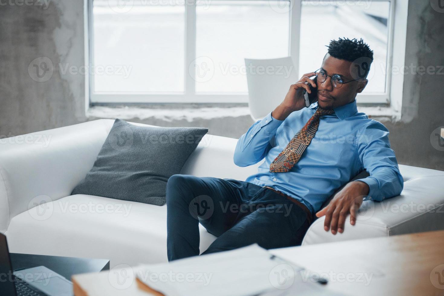 Foto von einem afroamerikanischen jungen Mann, der auf einer Couch sitzt und sich am Telefon unterhält?