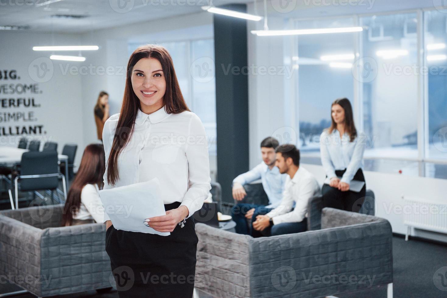 Womit kann ich Ihnen behilflich sein. Porträt des jungen Mädchens steht im Büro mit Mitarbeitern im Hintergrund foto