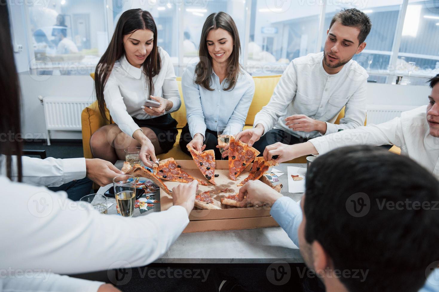 die Scheiben nehmen. Pizza essen. erfolgreiches Geschäft feiern. Junge Büroangestellte sitzen mit Alkohol am Tisch foto