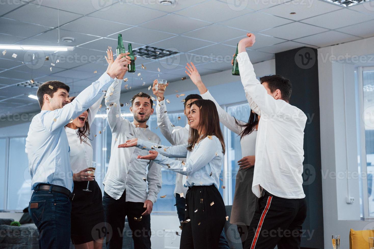 Konfetti in der Luft. Foto eines jungen Teams in klassischer Kleidung, das den Erfolg feiert, während es im modernen, gut beleuchteten Büro Getränke hält