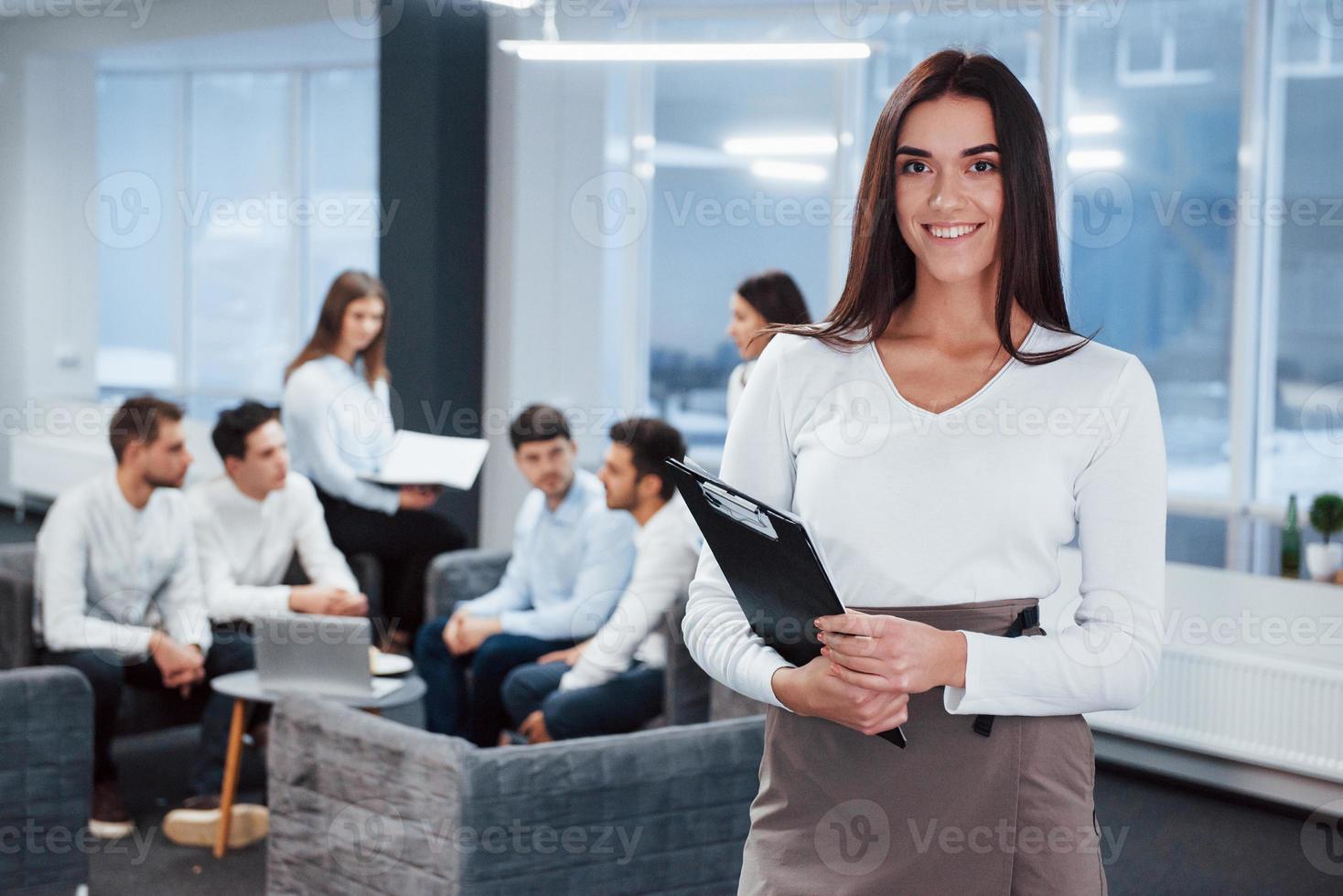 glücklicher Arbeiter. Porträt des jungen Mädchens steht im Büro mit Mitarbeitern im Hintergrund foto