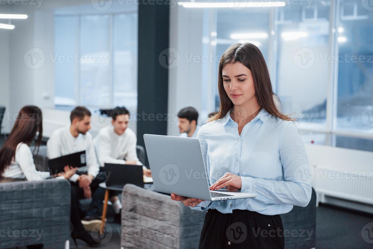 mit Laptop arbeiten. Porträt des jungen Mädchens steht im Büro mit Mitarbeitern im Hintergrund foto