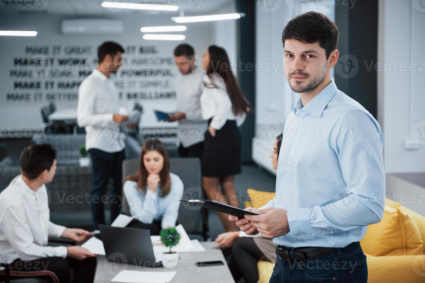 schönes Interieur. Porträt des jungen Mannes steht im Büro mit Mitarbeitern im Hintergrund foto