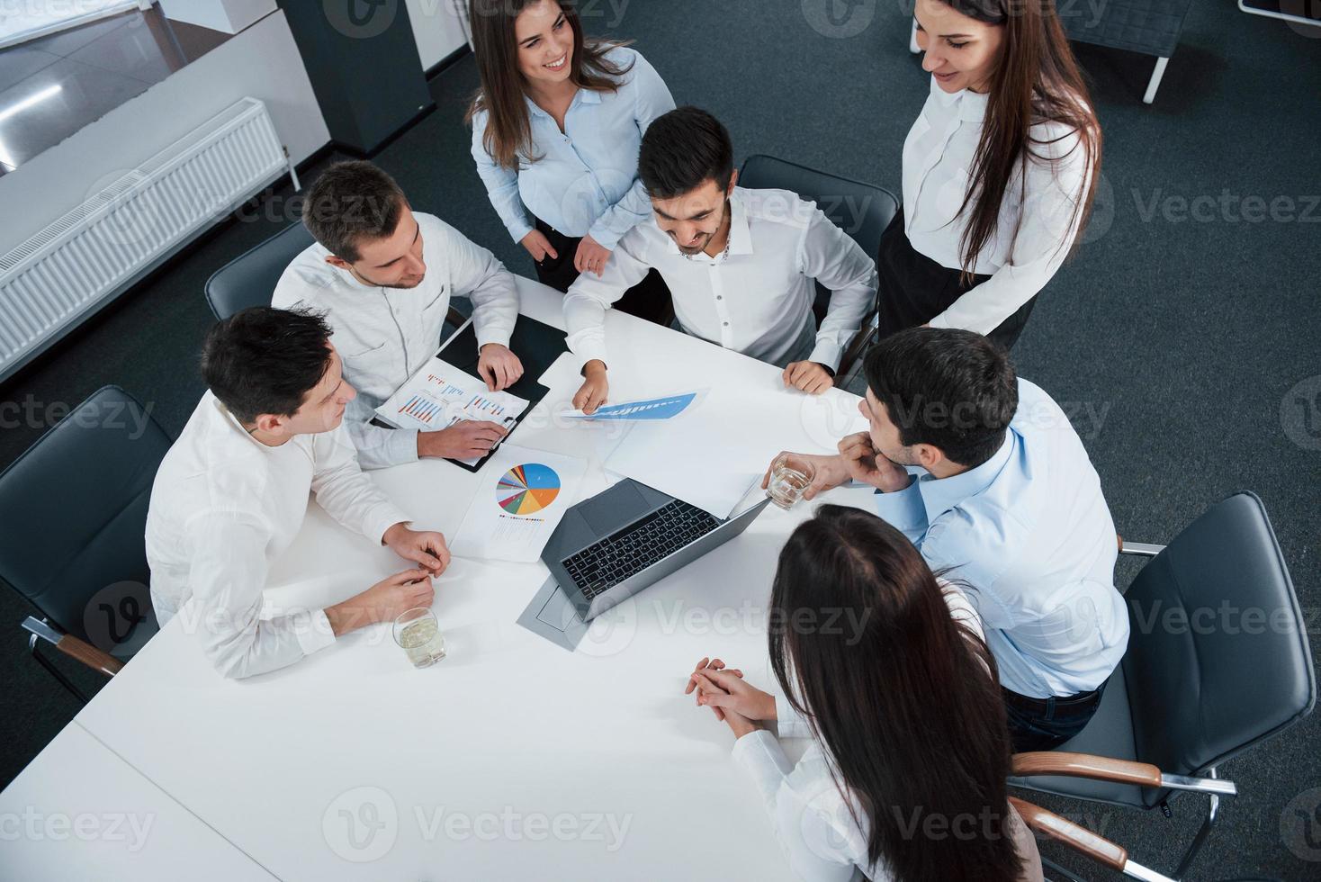 Kerl hält Getränk, Mädchen lächelnd. Draufsicht auf Büroangestellte in klassischer Kleidung, die mit Laptop und Dokumenten am Tisch sitzen foto