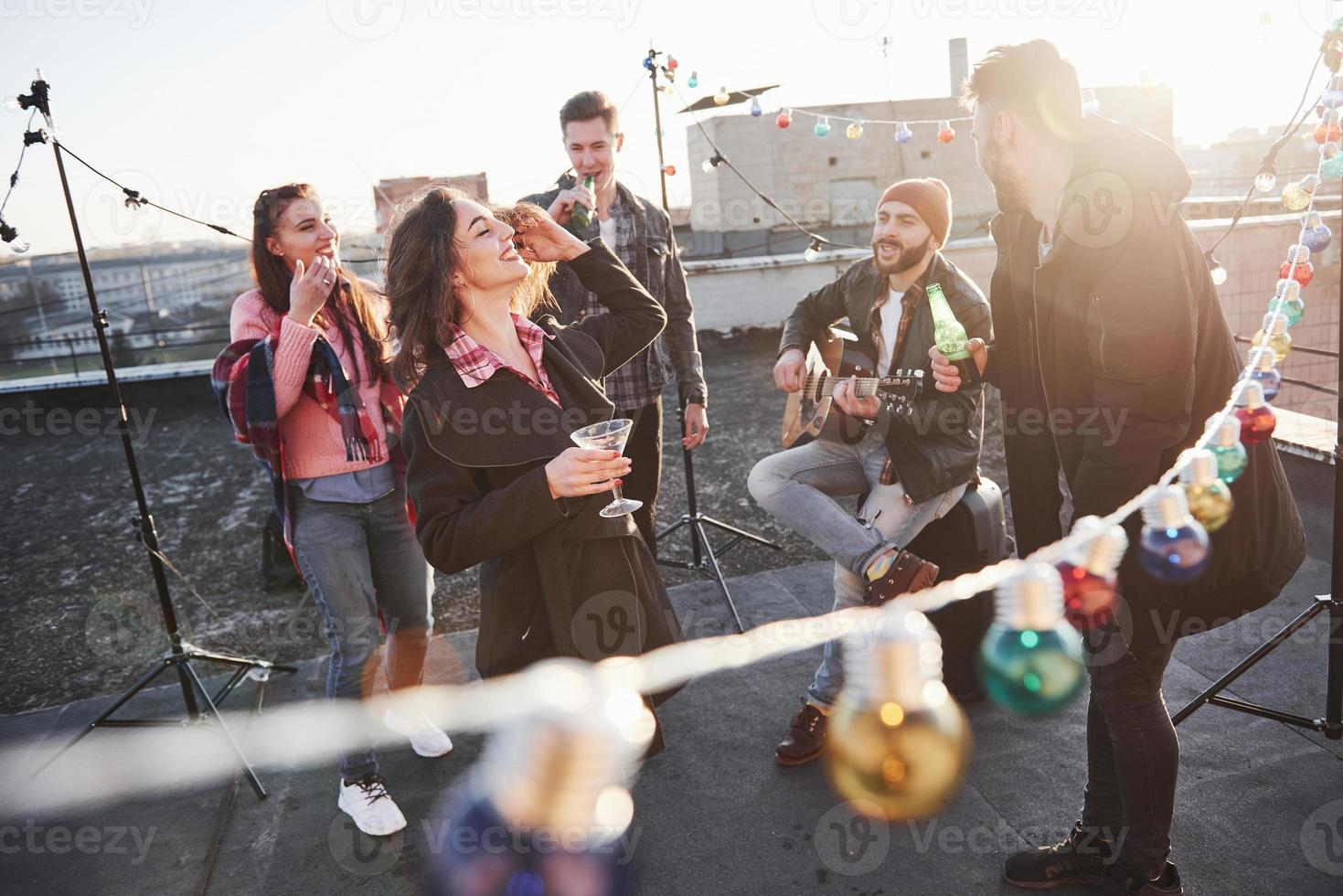 bärtiger Kerl weiß, wie man das Lächeln von den Leuten bekommt. Glühbirnen überall auf dem Dach, wo eine junge Gruppe von Freunden beschlossen hat, ihr Wochenende mit Gitarre und Alkohol zu verbringen? foto