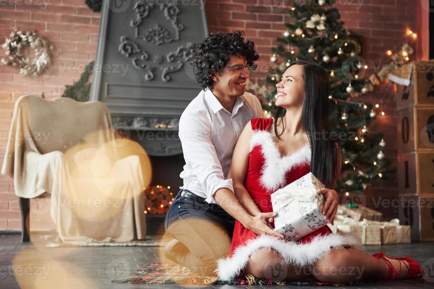 einander mit Liebe anschauen. Schönes Paar feiert Neujahr im dekorierten Raum mit Weihnachtsbaum und Kamin dahinter foto