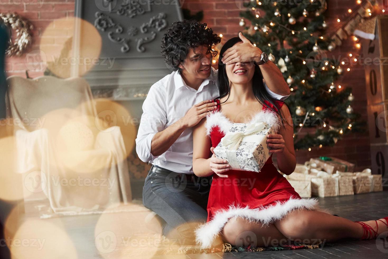 wunderschöner Moment. Mann macht Überraschung für seine Freundin. gibt Neujahrsgeschenk. junges Paar sitzt auf dem Boden des Zimmers des urlaubsgeschmückten Zimmers foto