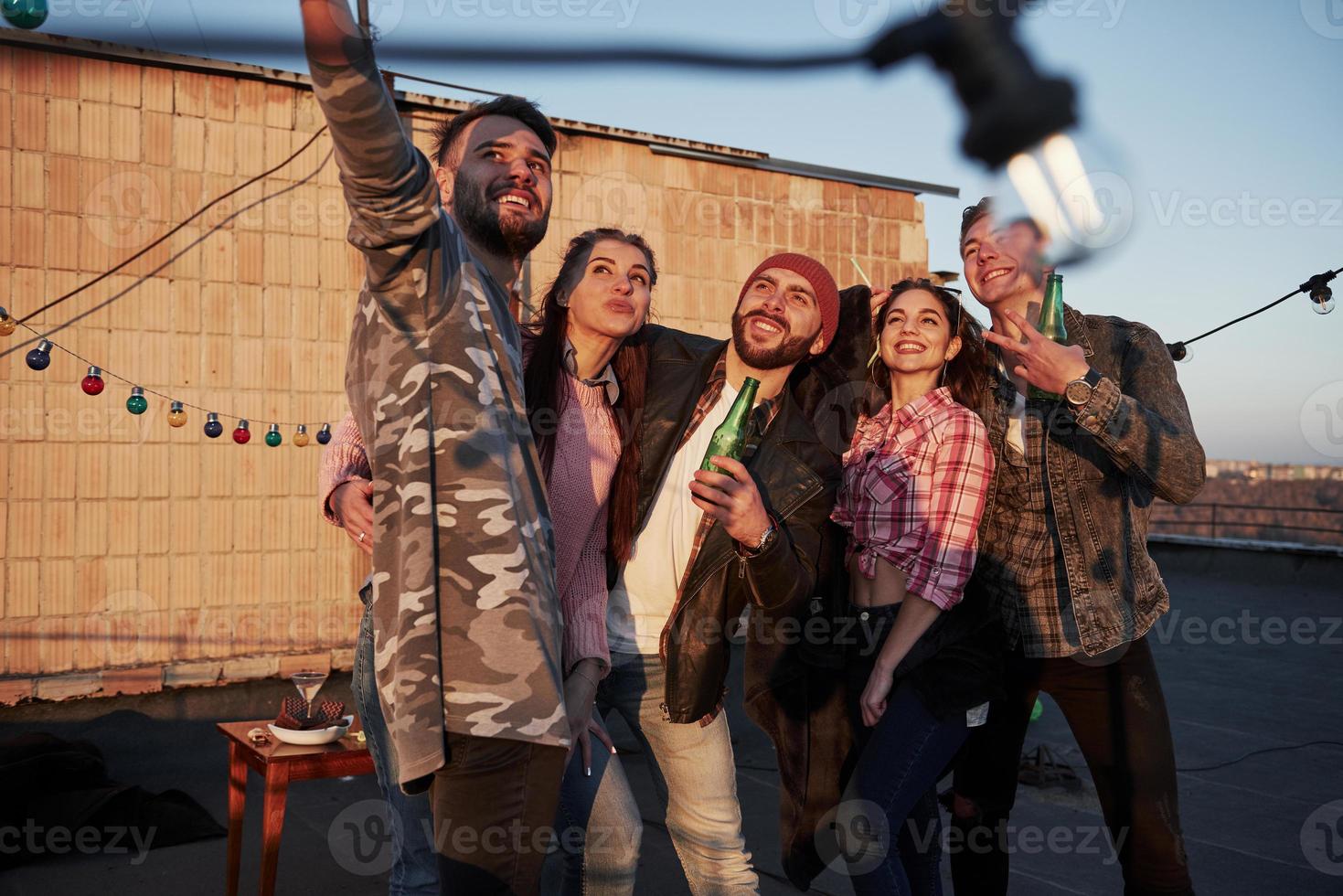 verschwommene Glühbirne vor der Kamera. Selfie-Zeit für eine Gruppe positiver Freunde steht auf dem Dach mit einer Wand dahinter foto