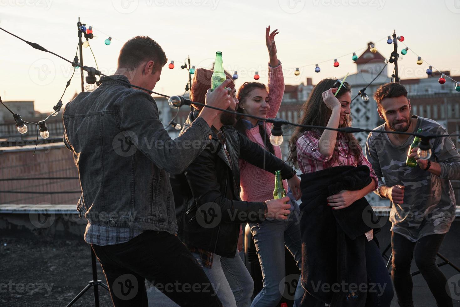 Alkohol in den Händen. Tanzparty auf dem Dach mit schönem Sonnenschein im Hintergrund. junge stylische Leute foto