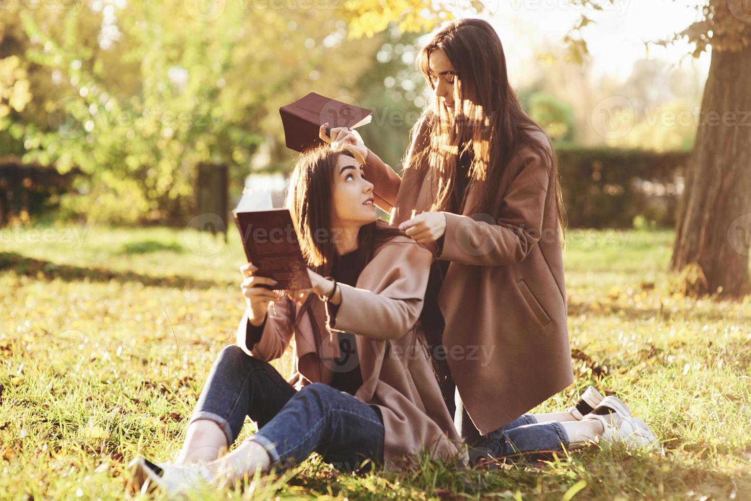 brünette Zwillingsmädchen, die mit braunen Büchern in den Händen auf dem Gras sitzen und sich ansehen, wenn eine von ihnen auf den Knien in der Nähe der Rückseite ihrer Schwester im Herbstpark auf verschwommenem Hintergrund steht foto