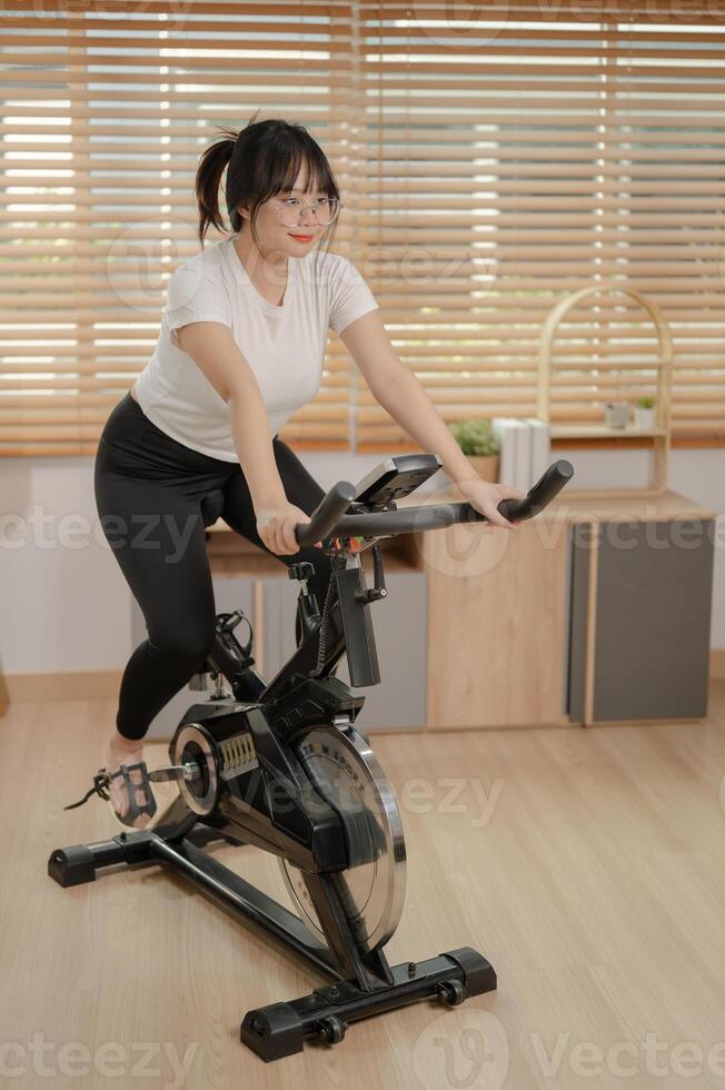 ein stark asiatisch Frau im Sportbekleidung ist ausüben auf ein Fitness Fahrrad, tun Innen- Cardio beim heim. foto