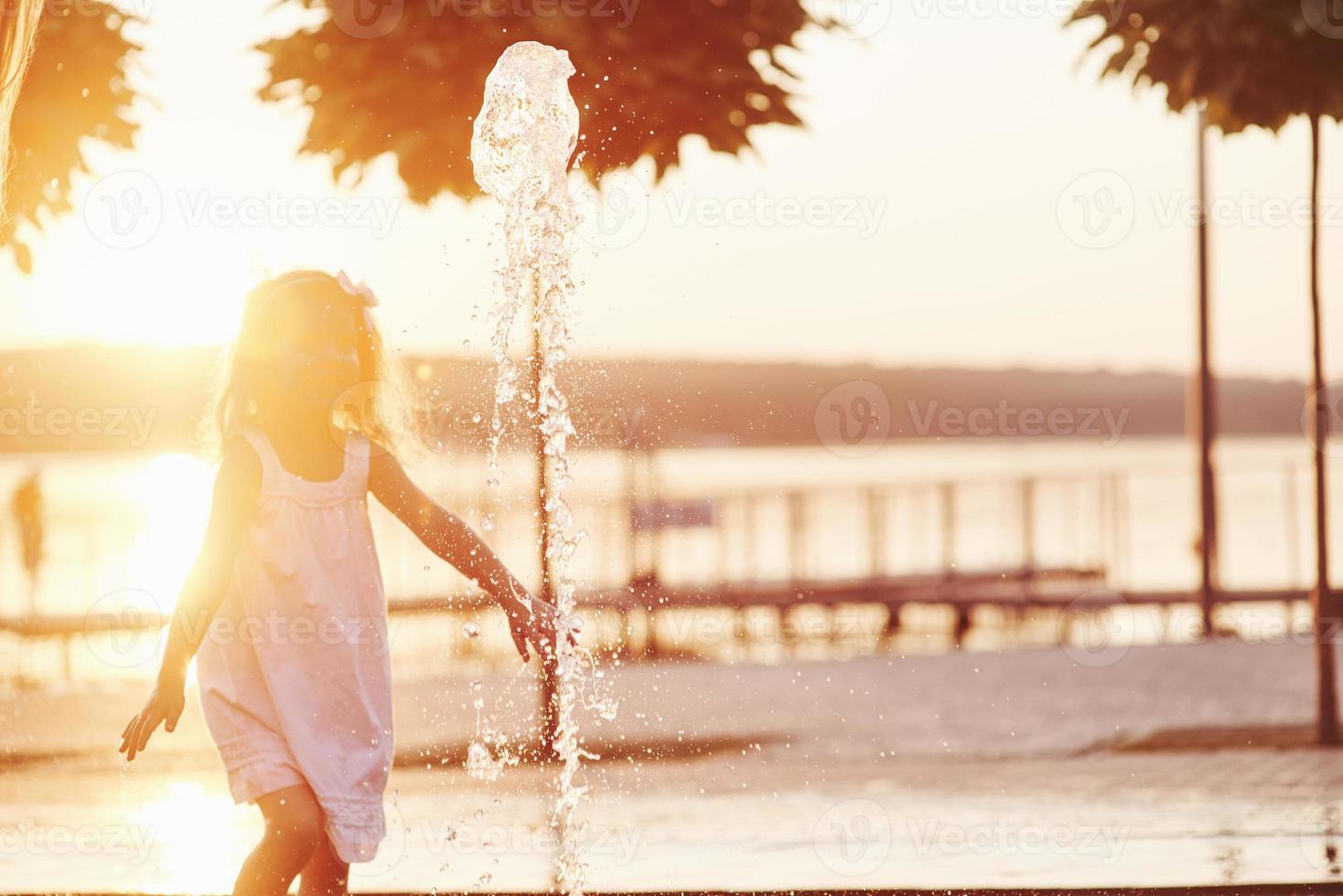 junges Mädchen spielt im Brunnen in der Sommerhitze und im See- und Waldhintergrund foto