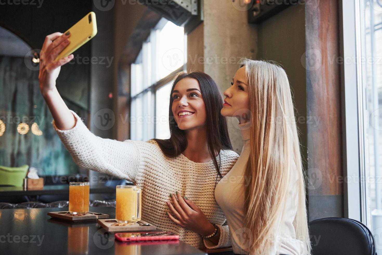 aufrichtige Brünette hat ein schönes Lächeln. Junge Freundinnen machen Selfie im Restaurant mit zwei gelben Getränken auf dem Tisch foto