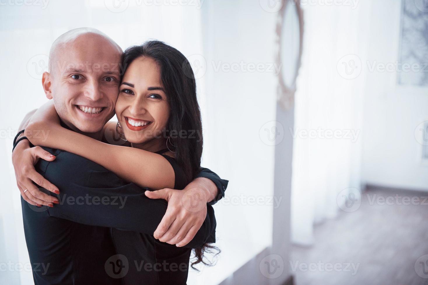 umarmen sich und lächeln. Porträt des glücklichen Paares drinnen. Glatzköpfiger und brünette Frau steht im weißen Raum foto