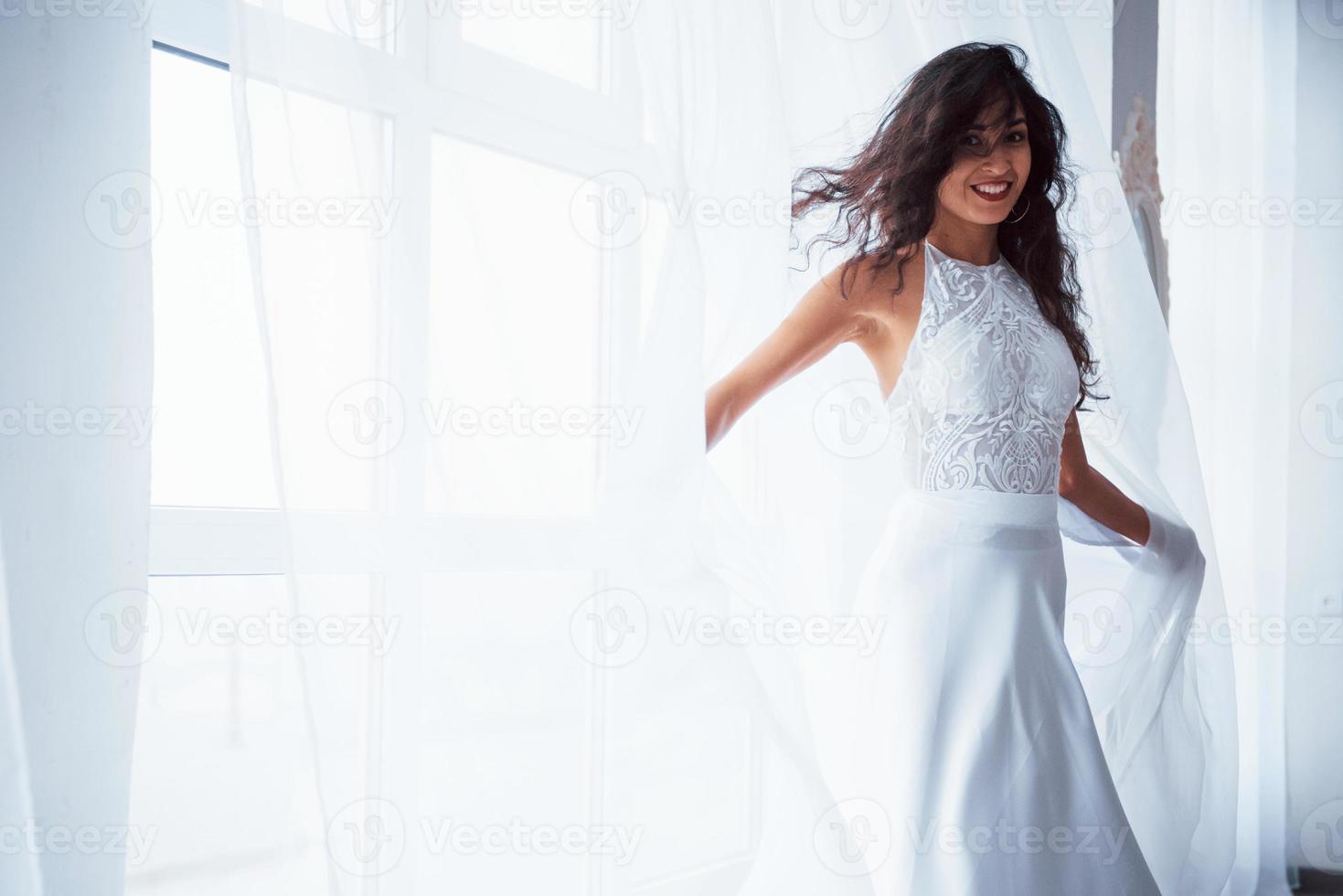 glänzendes Foto. Schöne Frau im weißen Kleid steht im weißen Raum mit Tageslicht durch die Fenster foto