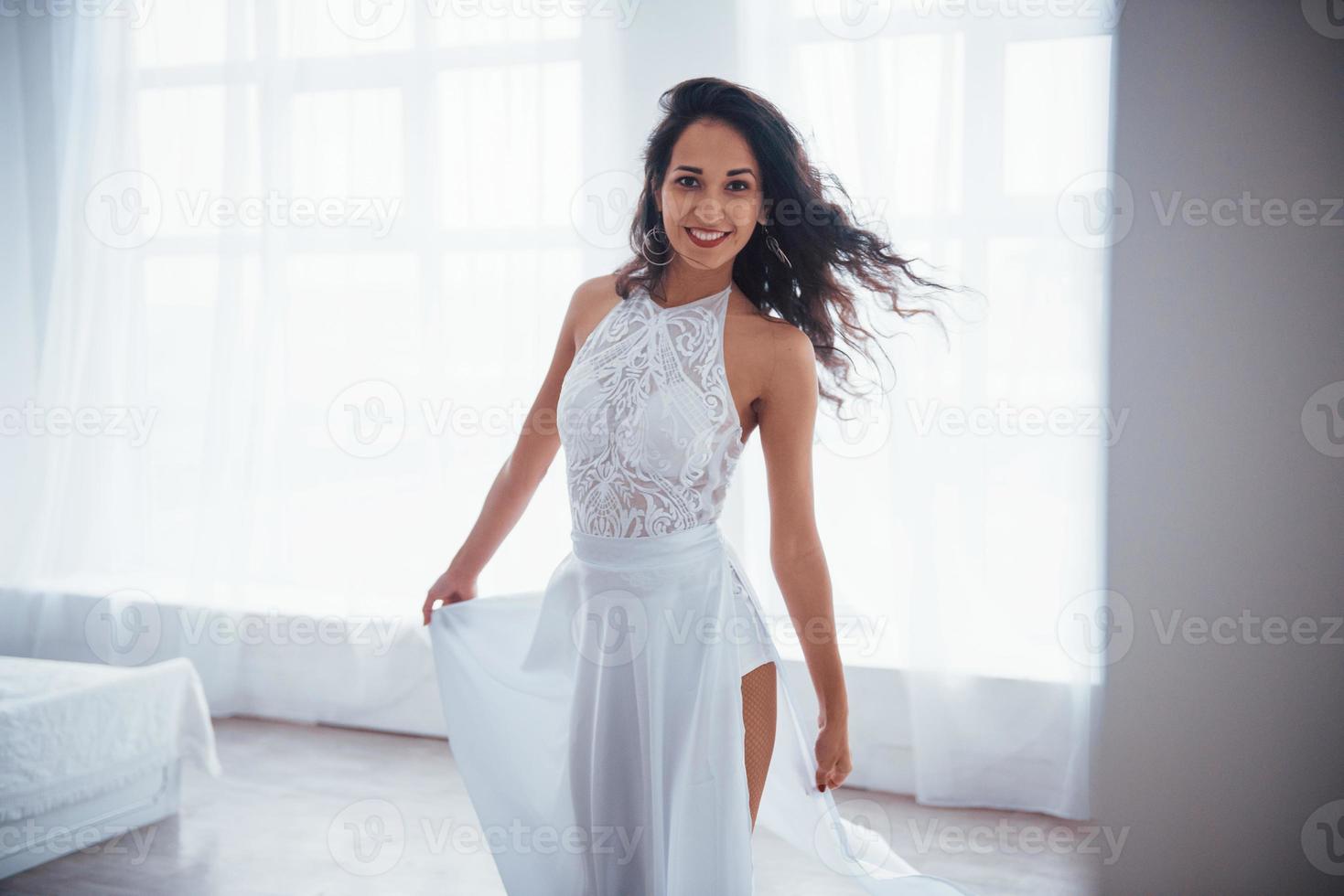 schöne Beleuchtung. Schöne Frau im weißen Kleid steht im weißen Raum mit Tageslicht durch die Fenster foto