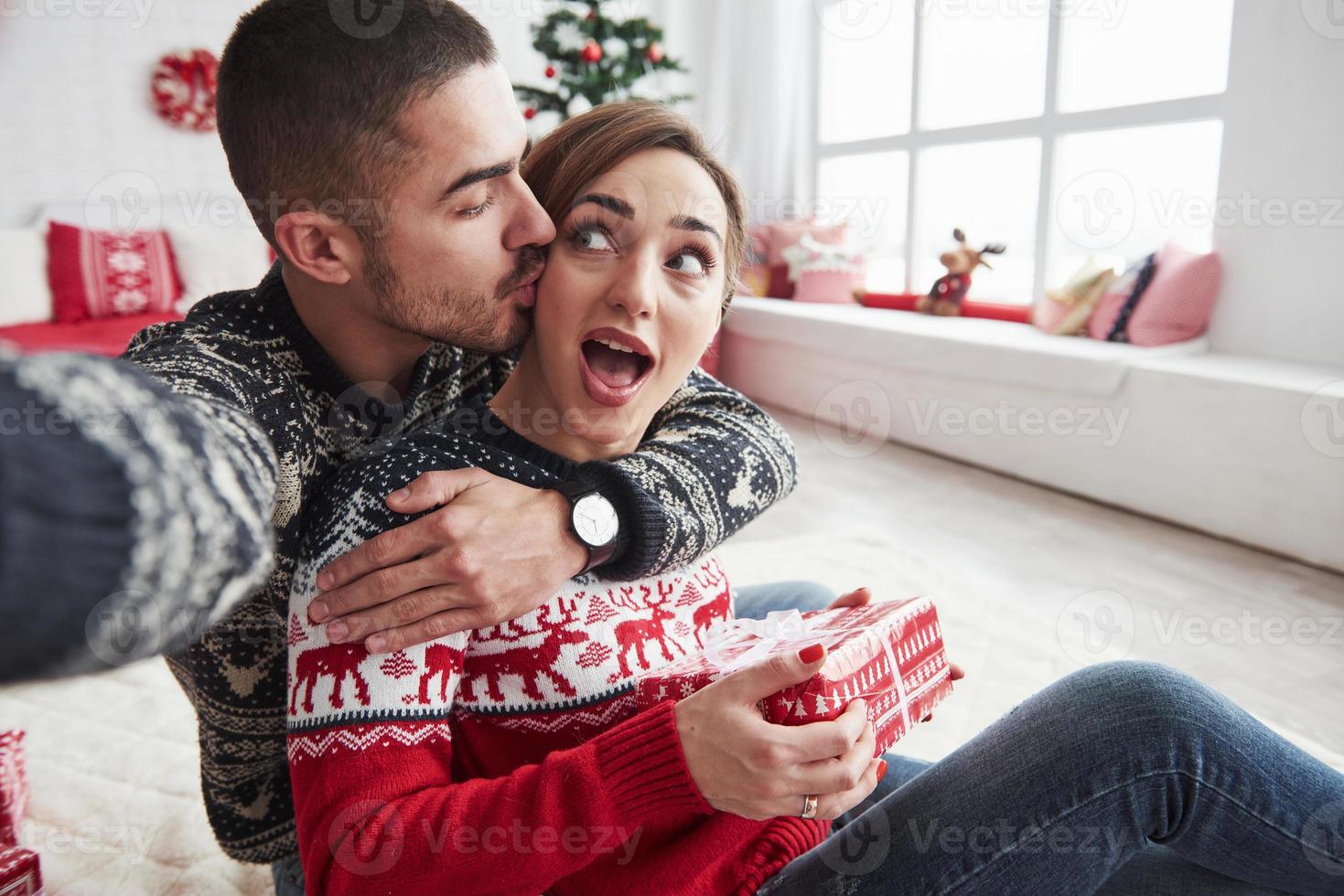 Frau geht auf ihren Freund zu, während sie das Geschenk hält. Mann, der ein Selfie von ihm und seiner Frau in Weihnachtskleidung macht und auf dem Boden eines dekorativen schönen Zimmers sitzt foto