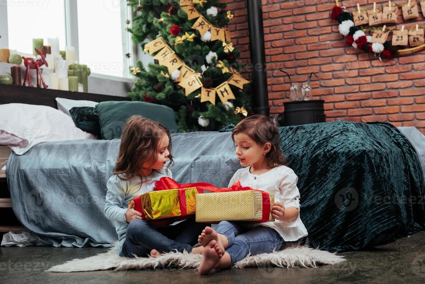 reden, während Sie bunte Kisten halten. Weihnachtsferien mit Geschenken für diese beiden Kinder, die drinnen im schönen Zimmer neben dem Bett sitzen foto