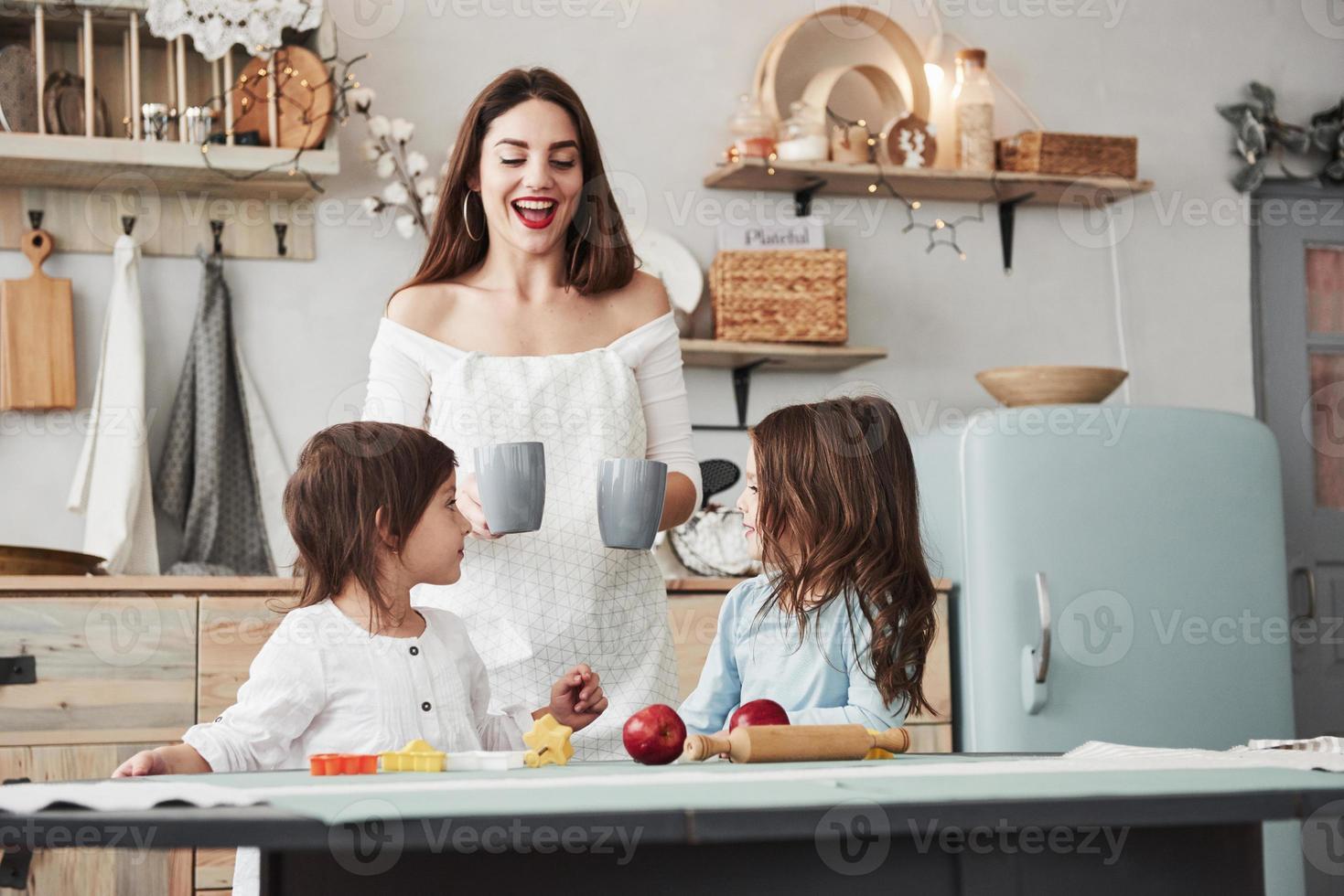 warten auf die Tassen. junge schöne frau gibt den kindern getränke, während sie mit spielzeug am tisch sitzen foto