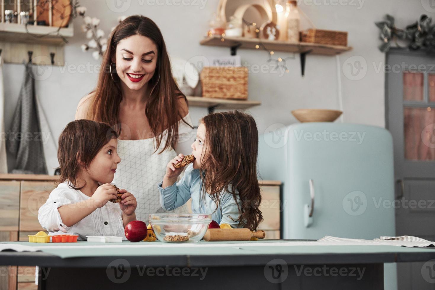 junge Mutter liebt es, wenn ihre Töchter zufrieden sind. junge schöne frau gibt die kekse, während sie mit spielzeug am tisch sitzen foto