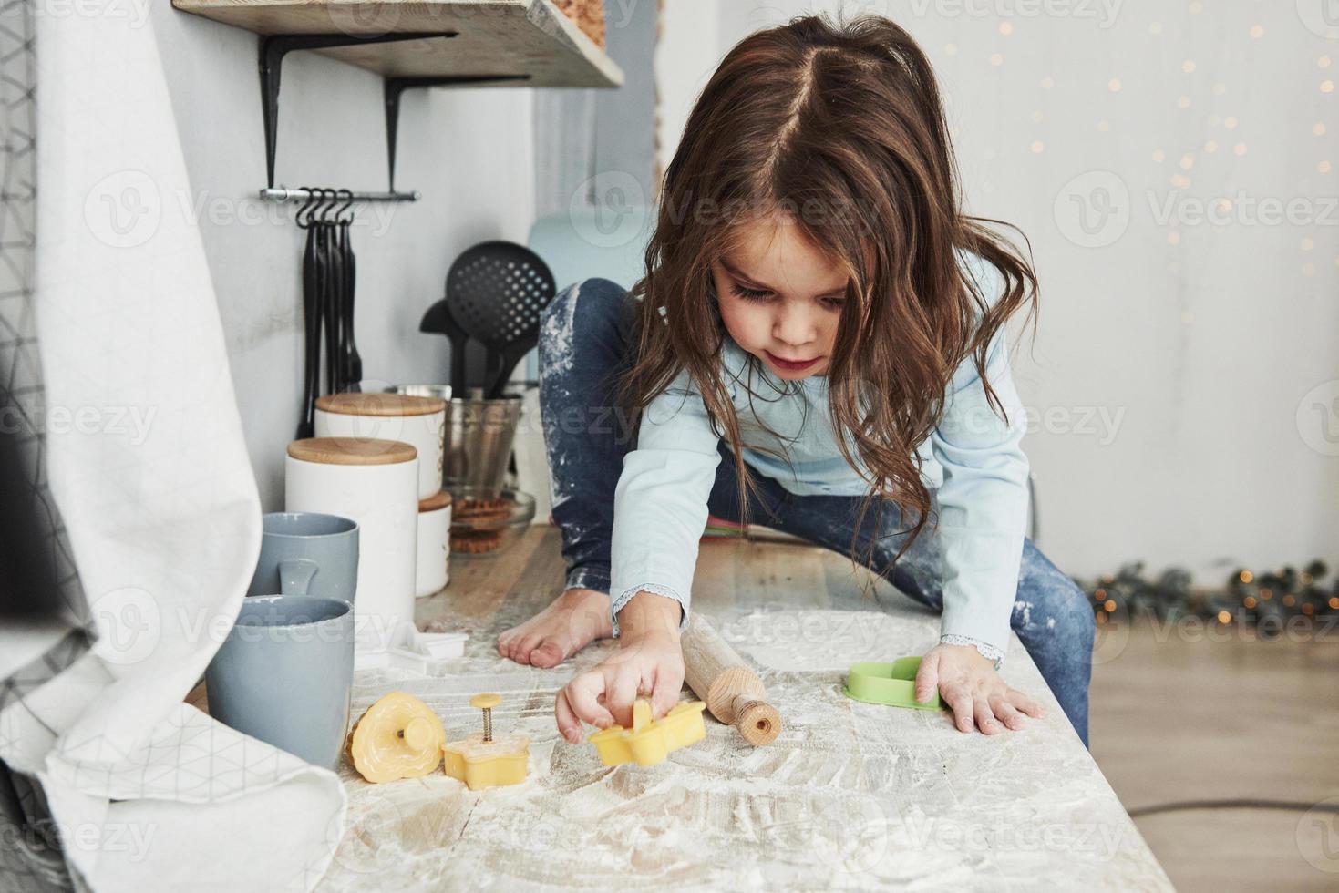 Vorderansicht. Foto eines hübschen kleinen Mädchens, das auf dem Küchentisch sitzt und mit Mehl spielt
