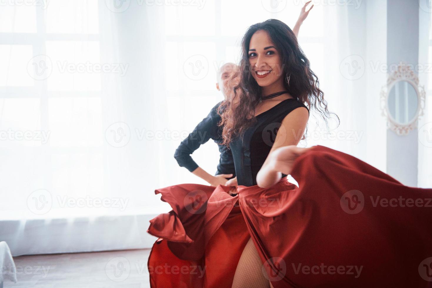 lächelt während des Prozesses. junge hübsche frau in rot-schwarzer kleidung tanzt mit glatzköpfigem mann im weißen raum foto