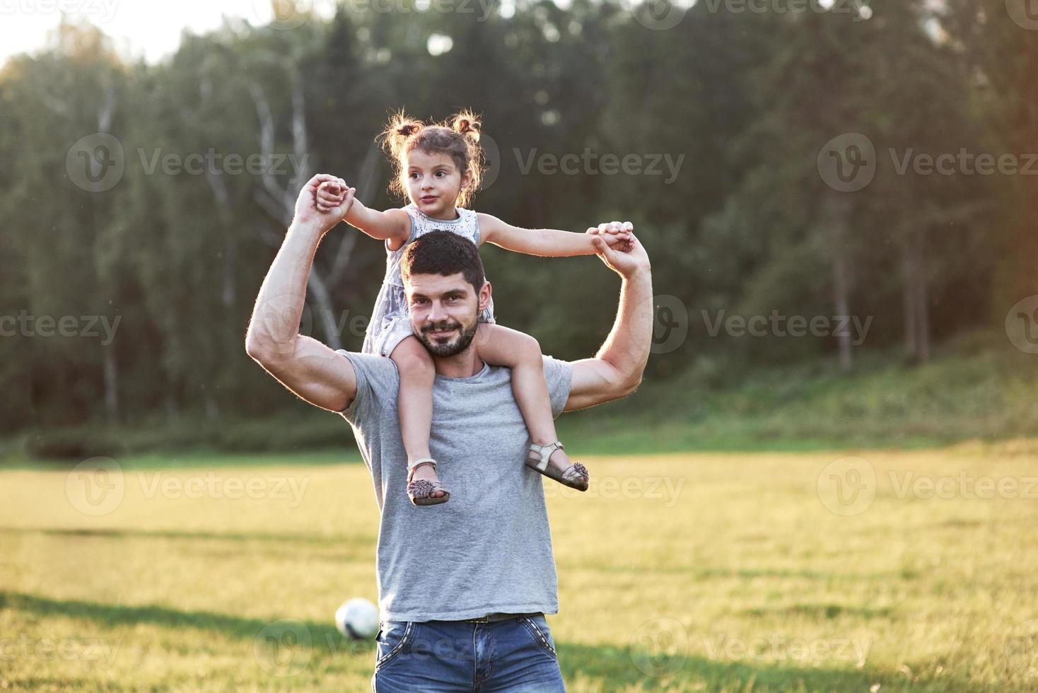 du wirst in Zukunft so aufwachsen wie ich, aber jetzt lass mich dich auf meine Schultern nehmen. Foto von Papa mit seiner Tochter bei schönem Gras und Wald im Hintergrund