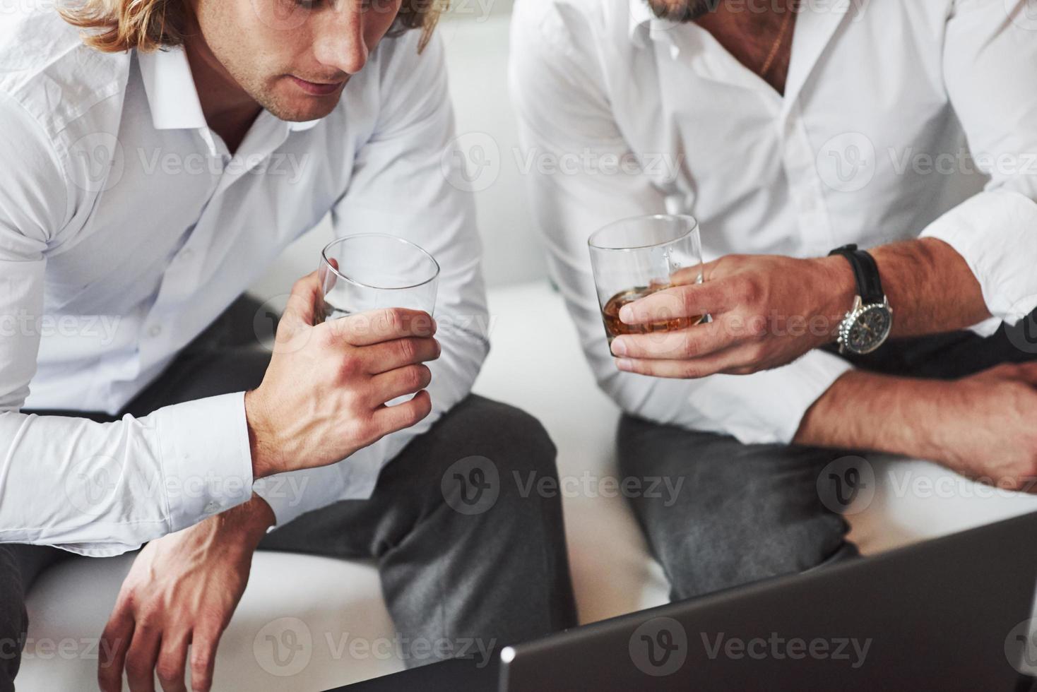 das Zeug suchen. Partikelansicht der beiden Geschäftsleute, die in der Nähe des Laptops sitzen und Whiskyglas halten foto