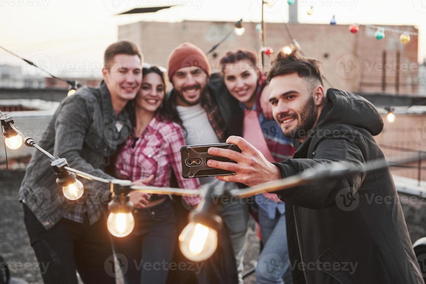 Konzentrieren Sie sich auf das Smartphone. Gruppe junger fröhlicher Freunde, die Spaß haben, sich umarmen und Selfies auf dem Dach mit dekorativen Glühbirnen machen foto