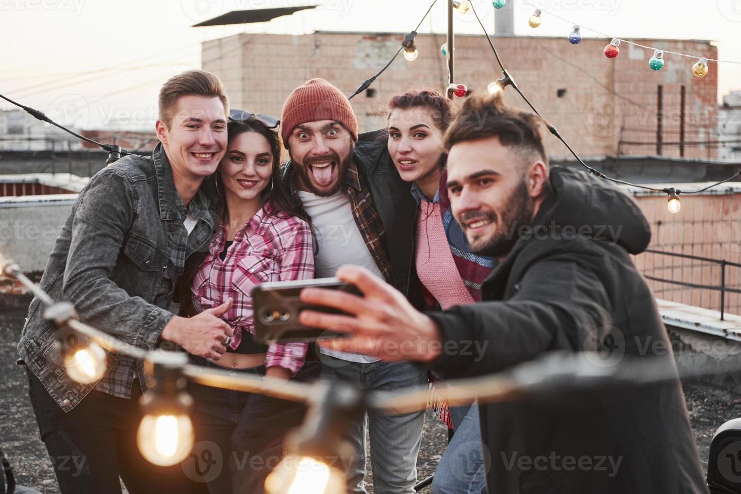 Kerl in der Mitte macht lustiges Gesicht. Gruppe junger fröhlicher Freunde, die Spaß haben, sich umarmen und Selfies auf dem Dach mit dekorativen Glühbirnen machen foto