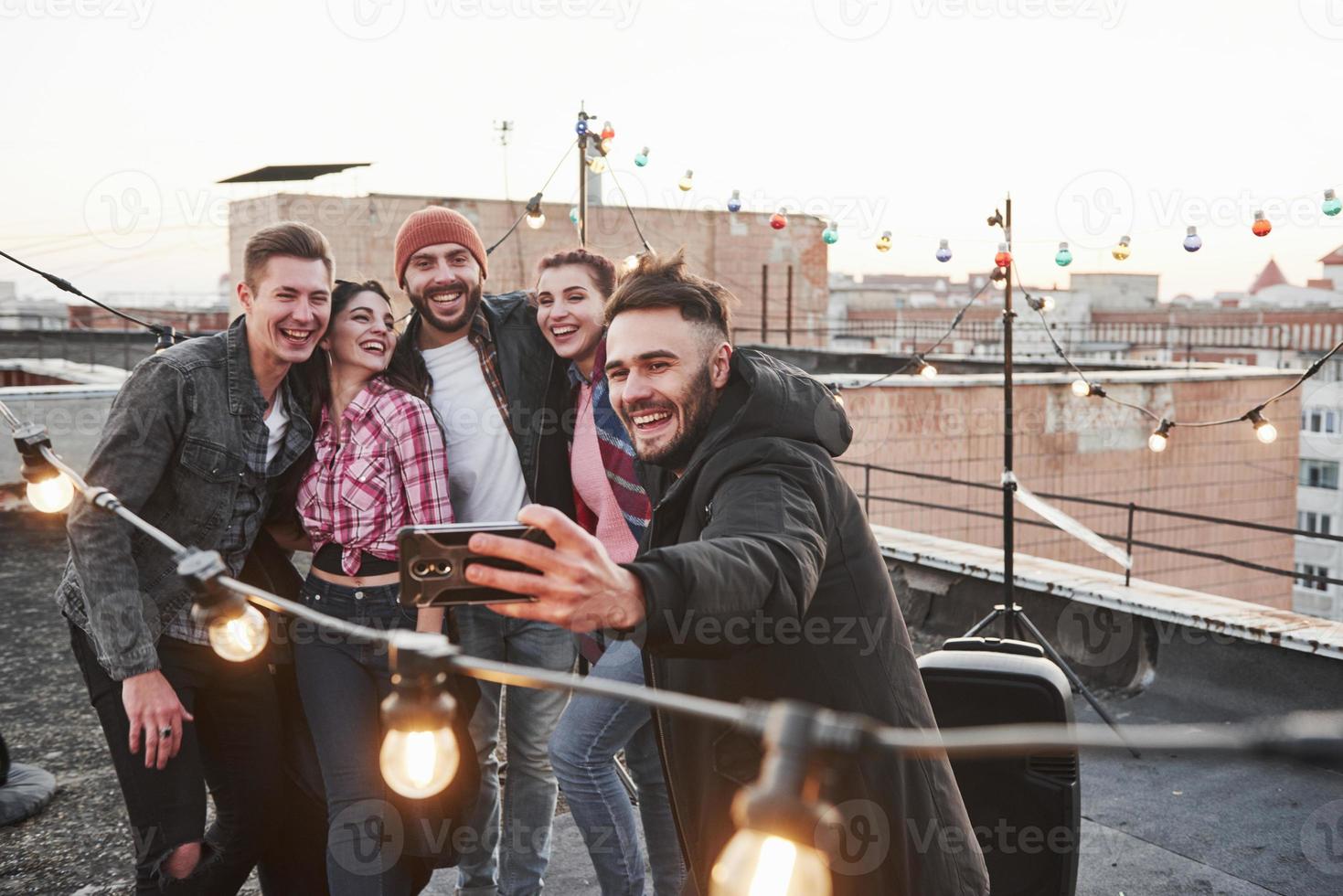 gute Laune. Gruppe junger fröhlicher Freunde, die Spaß haben, sich umarmen und Selfies auf dem Dach mit dekorativen Glühbirnen machen foto