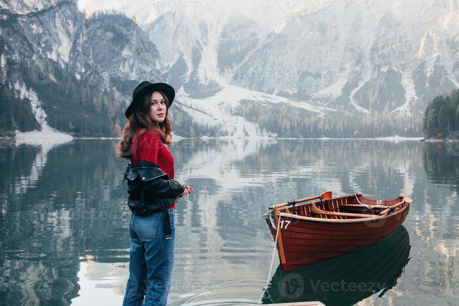Mädchen, das nach hinten schaut. hervorragende Aussicht. Frau mit schwarzem Hut genießt majestätische Berglandschaft in der Nähe des Sees mit Boot foto