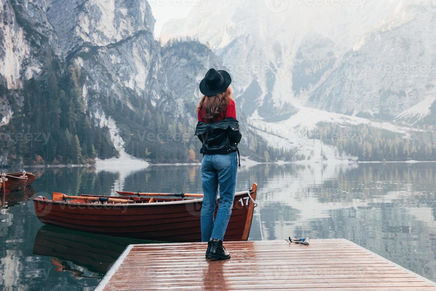 Foto von hinten. Frau mit schwarzem Hut genießt majestätische Berglandschaft in der Nähe des Sees mit Booten