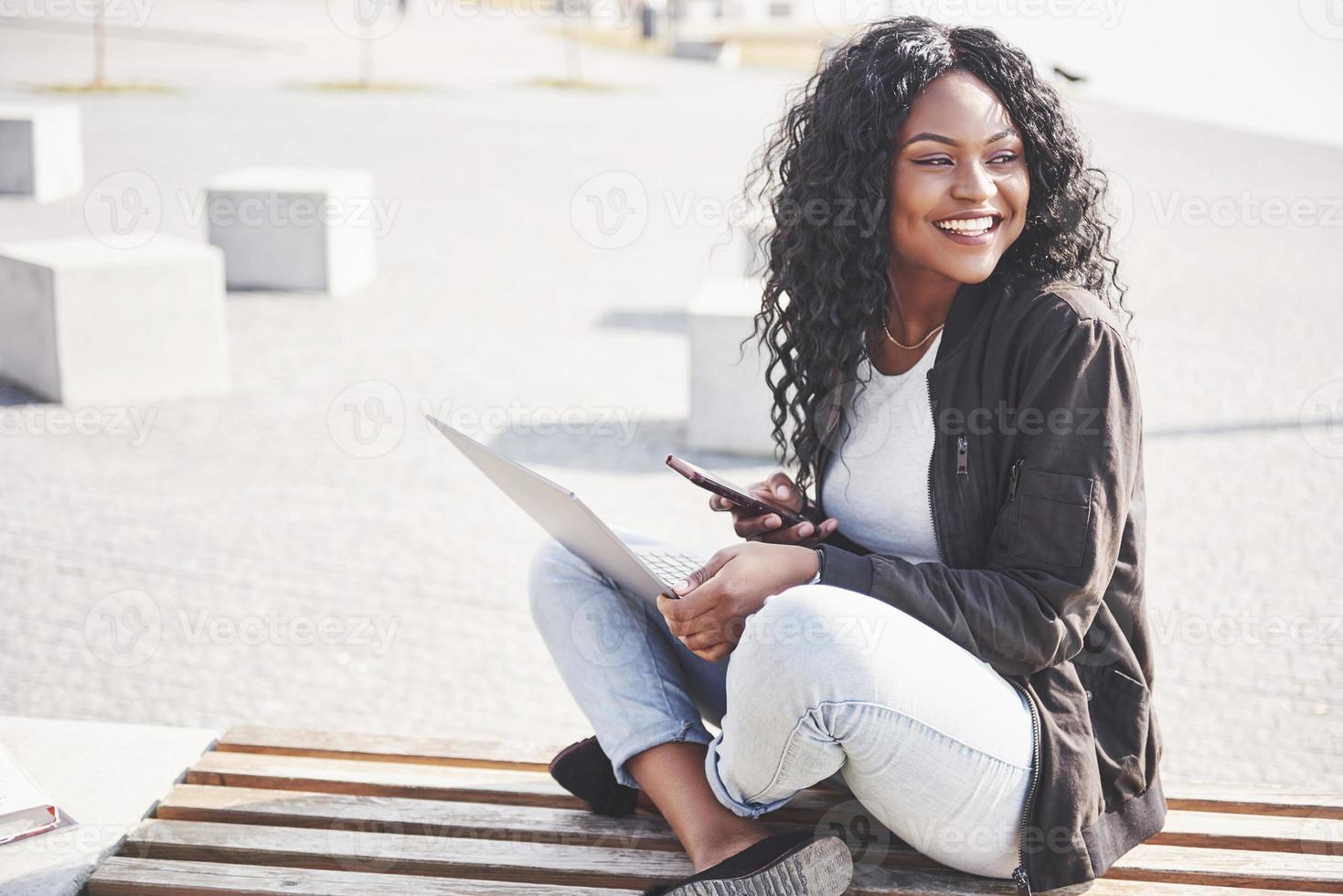 junge Frau auf der Straße, die am Laptop arbeitet und mit dem Handy spricht foto