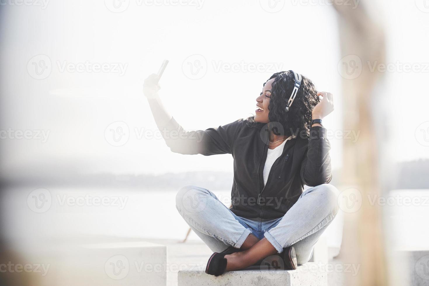 Porträt eines schönen jungen hübschen afroamerikanischen Mädchens, das am Strand oder See sitzt und Musik in ihren Kopfhörern hört foto