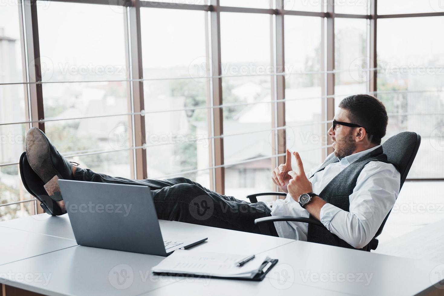 Schöner junger Geschäftsmann mit Brille, der seine Beine auf dem Tisch hält und einen Laptop im Büro betrachtet foto