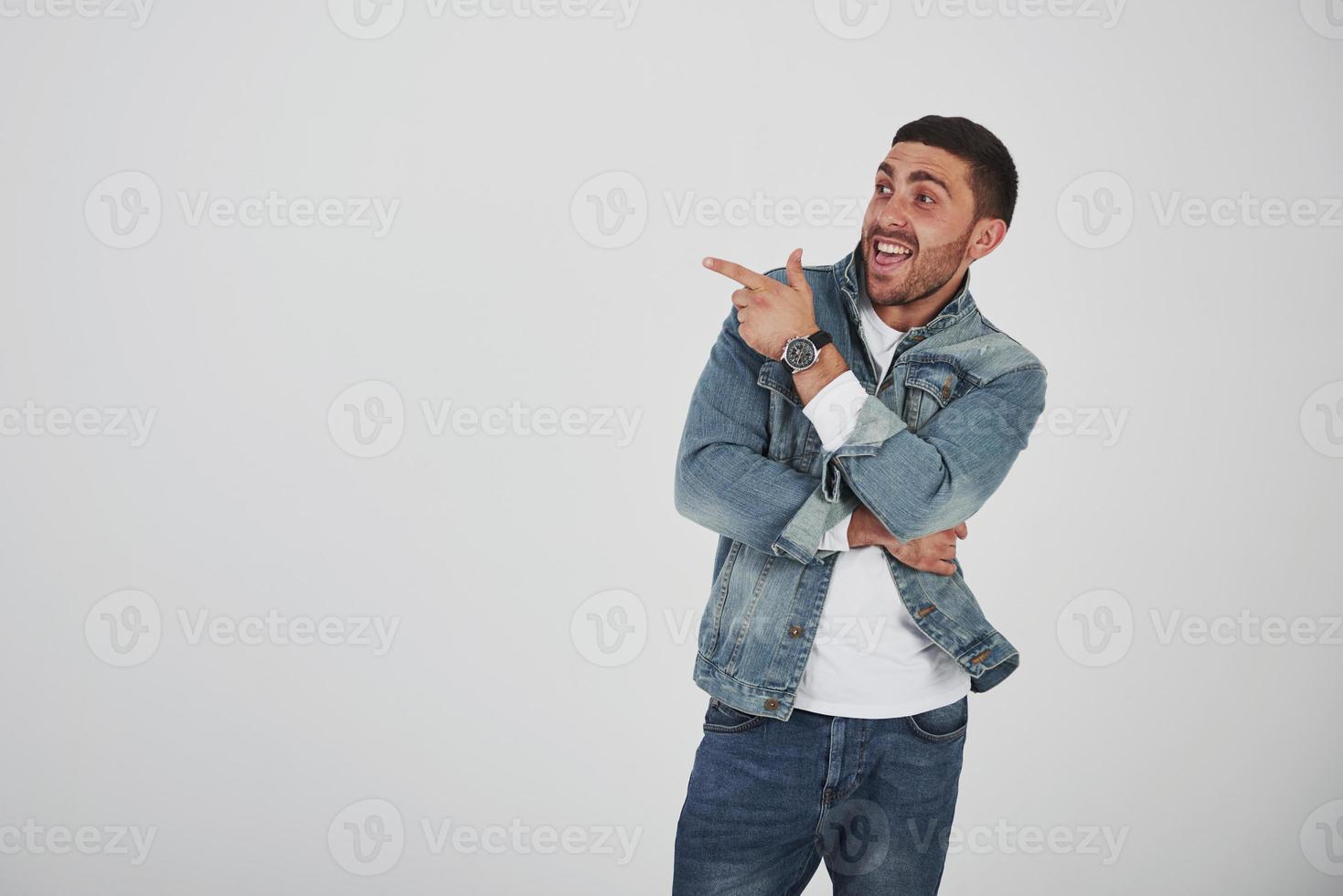 Glücklicher aufgeregter bärtiger Mann mit Brille, der mit einem Lächeln in die Kamera schaut und mit zwei Fingern auf weißem Hintergrund auf den Kopierraum zeigt foto