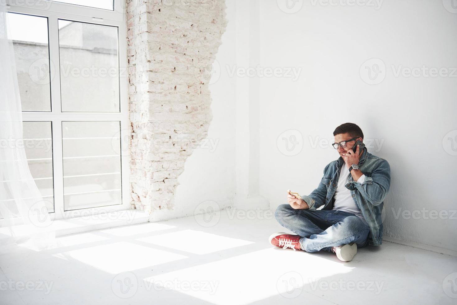 Ein Mann in Freizeitkleidung sitzt zu Hause in einer leeren Wohnung, hält eine Kreditkarte und ruft an. Vielleicht ist er ein neuer Bewohner und hat noch keine Möbel gekauft. foto