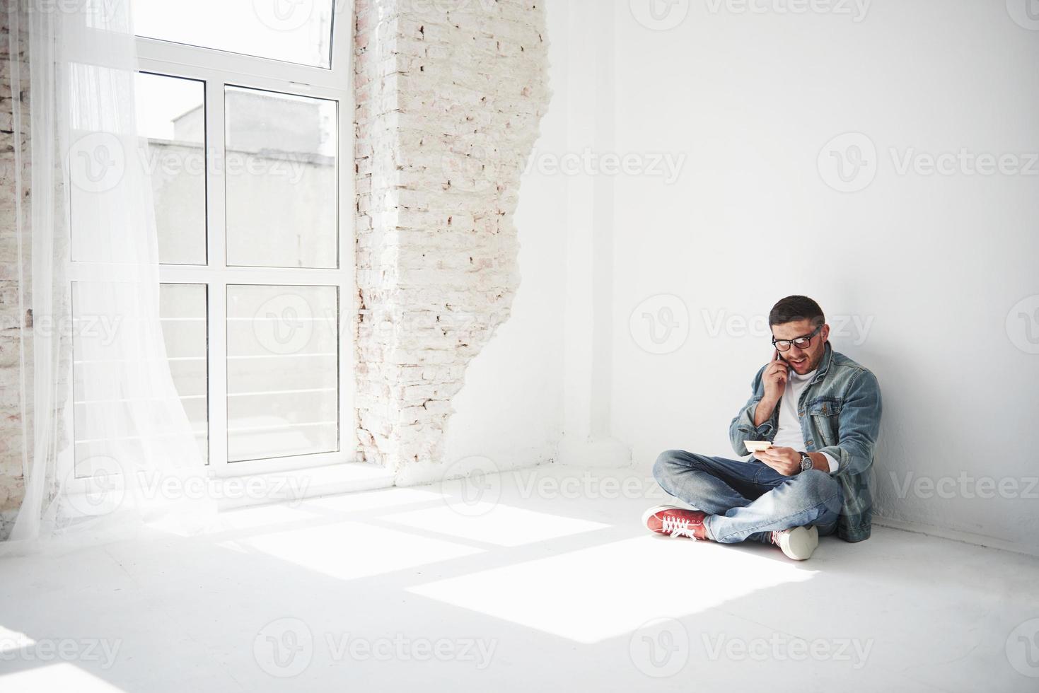 ein mann in lässiger kleidung sitzt zu hause in einer leeren wohnung und telefoniert. vielleicht wurde er ausgeraubt, oder sie nahmen den ganzen Besitz für Schulden ein foto