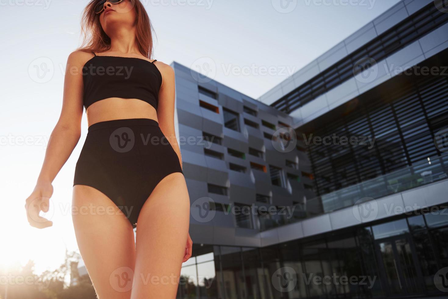 sexy junge Frau mit Sonnenbrille und schwarzem Badeanzug steht während des Sonnenuntergangs auf dem Hintergrund der Stadtgebäude foto