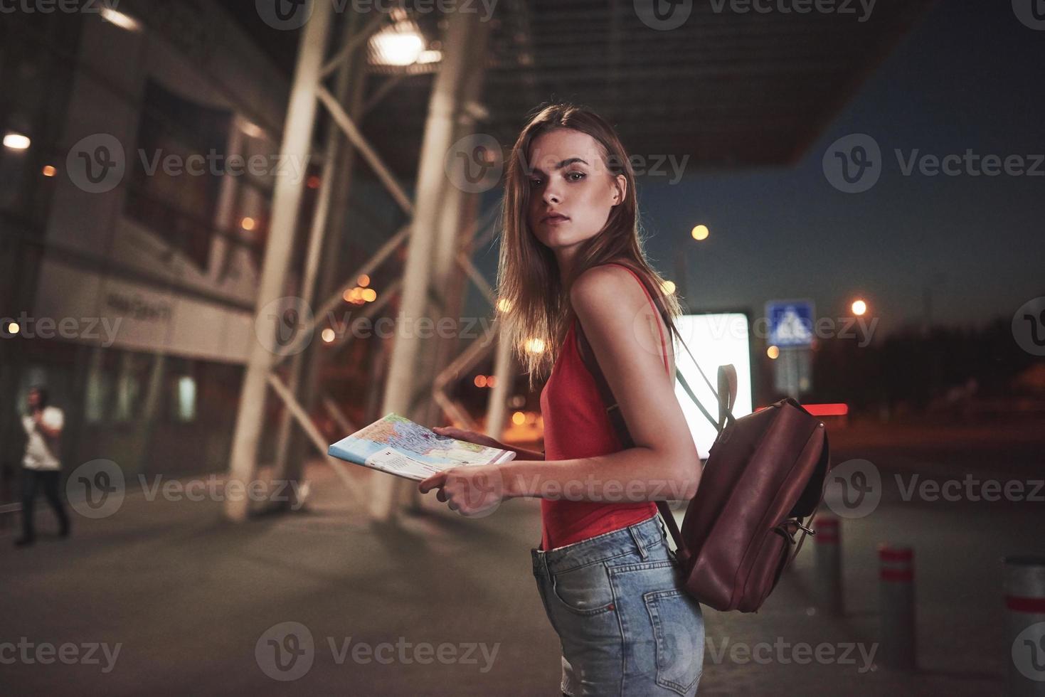 Ein junges schönes Mädchen mit einem Rucksack hinter den Schultern steht an einem warmen Sommerabend auf der Straße in der Nähe eines Flughafens oder eines Bahnhofs. sie ist gerade angekommen und wartet auf ein Taxi oder ihre Freunde foto