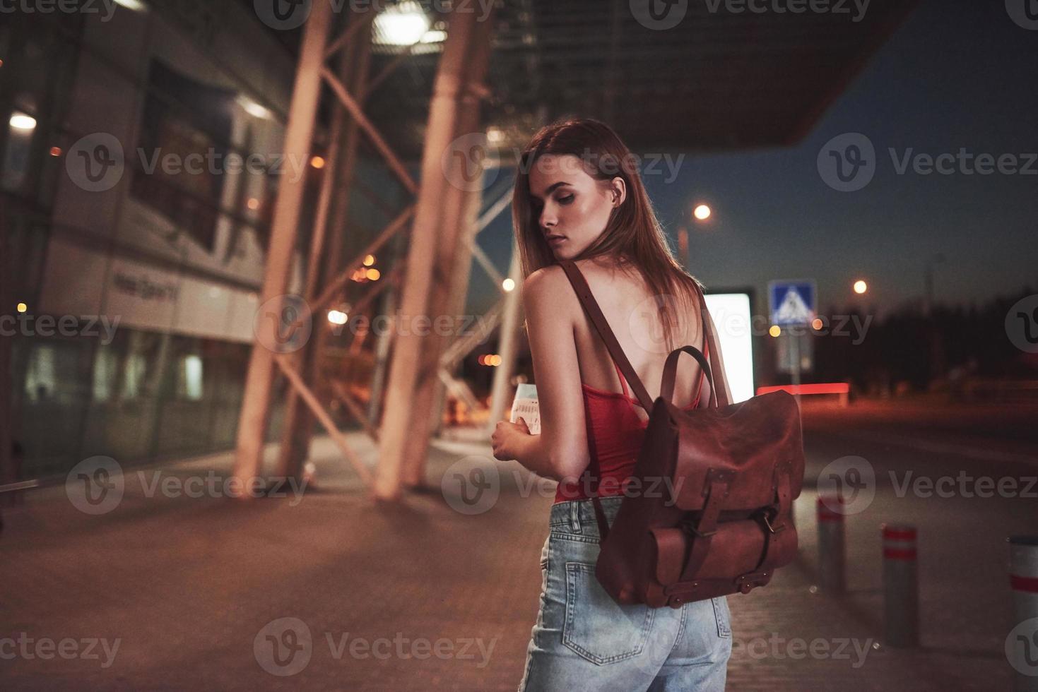 Ein junges schönes Mädchen mit einem Rucksack hinter den Schultern steht an einem warmen Sommerabend auf der Straße in der Nähe eines Flughafens oder eines Bahnhofs. sie ist gerade angekommen und wartet auf ein Taxi oder ihre Freunde foto