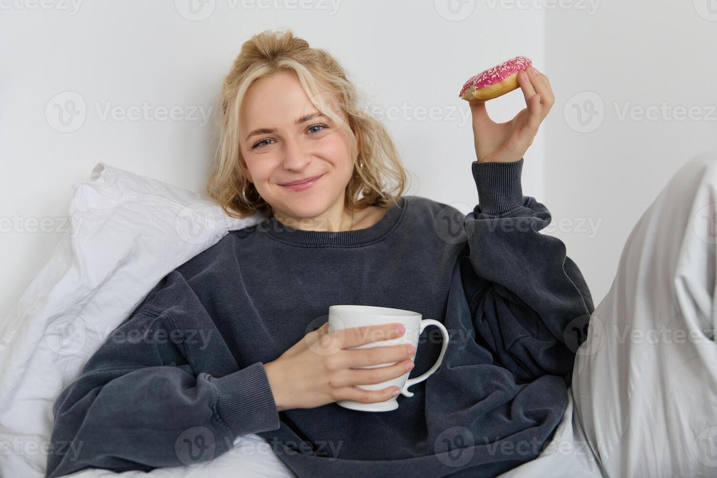 Lebensstil und Menschen Konzept. glücklich blond Frau Lügen im Bett mit Essen, Trinken Tee und Essen Krapfen, ruhen beim heim, Ausgaben Zeit allein, Aufpassen Fernseher Show foto