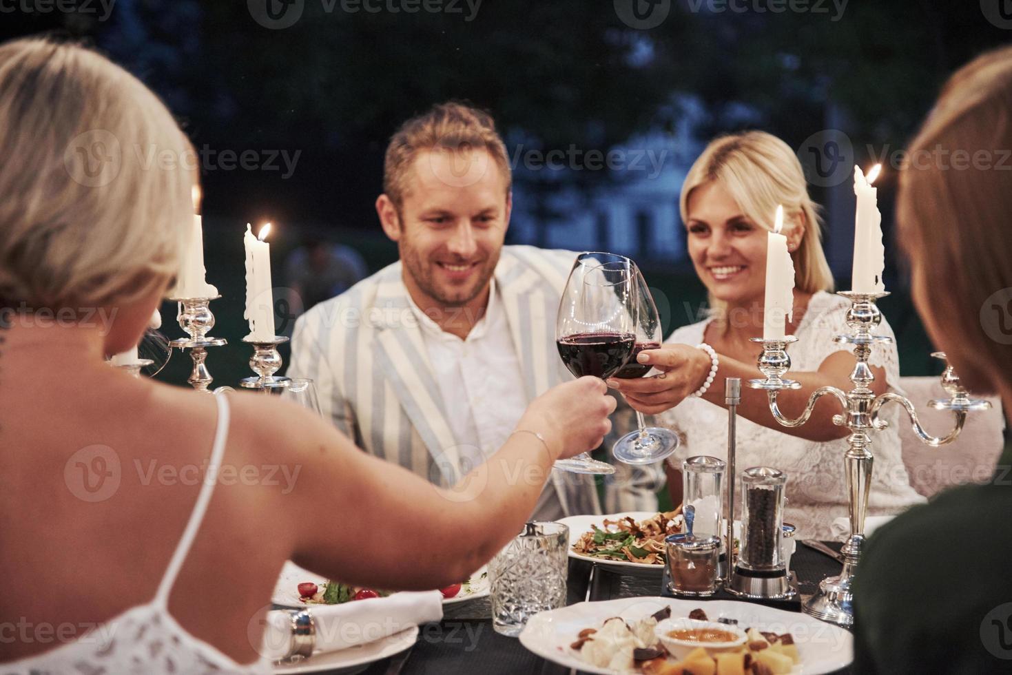 mit Wein auf die Gläser klopfen. Gruppe von Freunden in der eleganten Kleidung haben ein luxuriöses Abendessen foto