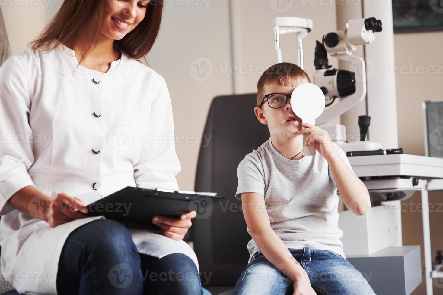 sieht aus, als ob die Frau ihren Job liebt. Frau mit Dokumenten testet die Sehschärfe eines kleinen Jungen foto