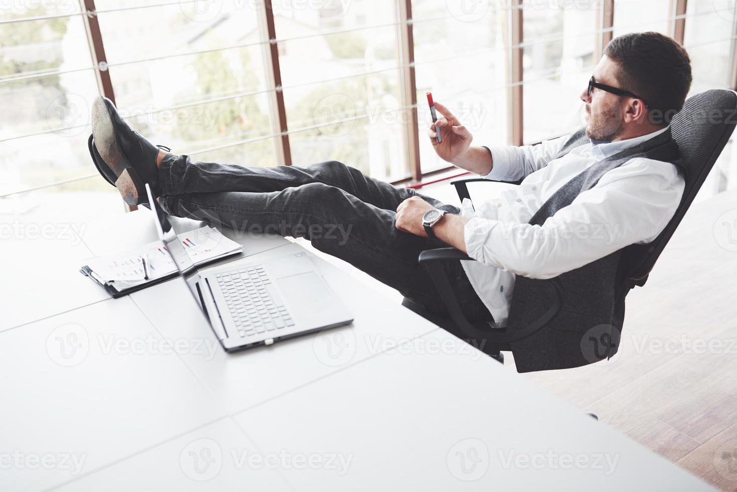 junger Geschäftsmann, der sich beim Sitzen auf einem Stuhl ausruht und im Reinraum die Beine auf den Tisch legt. durch die Fenster gucken foto