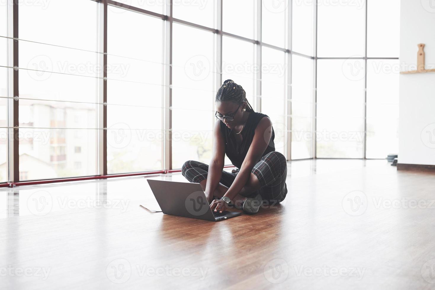 schönes afroamerikanisches Mädchen, das auf dem Holzboden sitzt und mit dem Laptop arbeitet foto